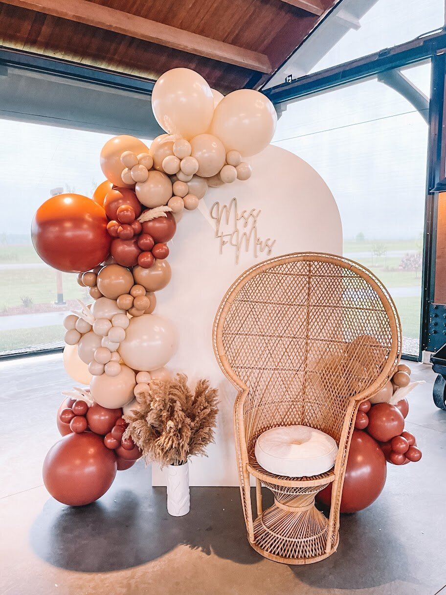 Boho bridal shower decorations. A white wooden arch with neutral balloons attached, pampas grass, and a peacock chair.