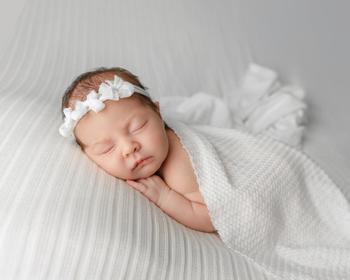 Newborn baby girl swaddled in soft white textures with a delicate bow headband, posed on a ribbed blanket during her studio session. Captured by Inland Empire newborn photographer Kaitlyn Dawn Photography.