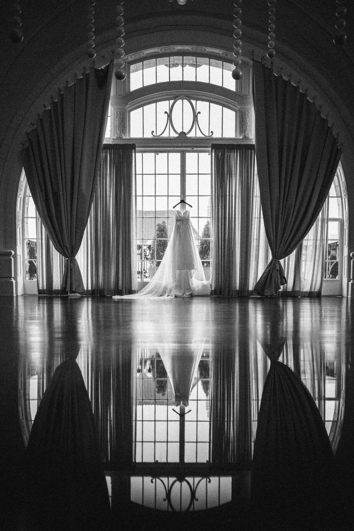 Black and white image of a wedding dress hanging in front of a large arched window with sheer curtains, framed by tall drapes. The dress and window are reflected in the polished ballroom floor, creating a symmetrical composition.