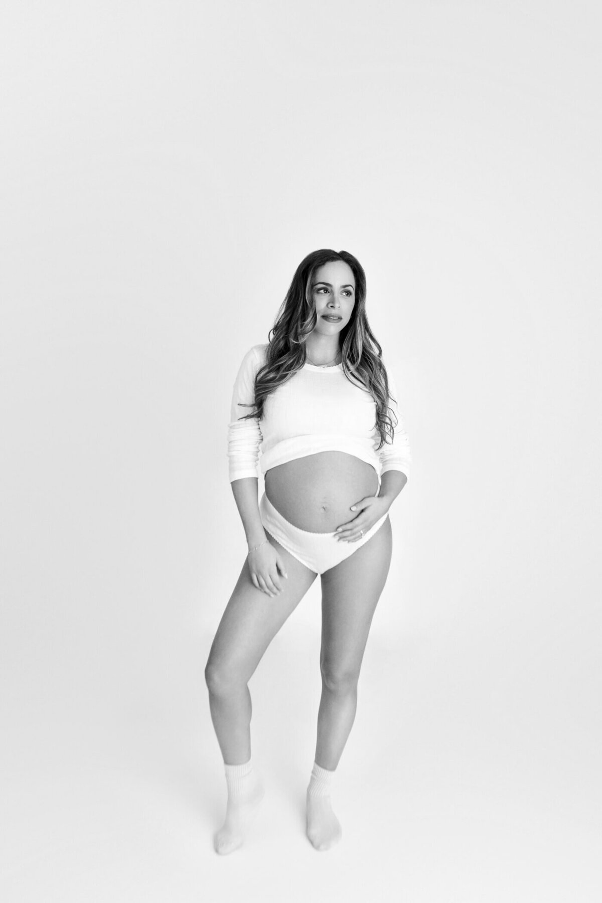 A black-and-white maternity portrait of a pregnant woman standing confidently against a white background. She wears a cropped white long-sleeve top, white underwear, and socks, revealing her baby bump. With one hand on her hip and the other resting on her belly, she gazes slightly off to the side with a serene expression. The minimalist setting highlights the simplicity and beauty of the moment.