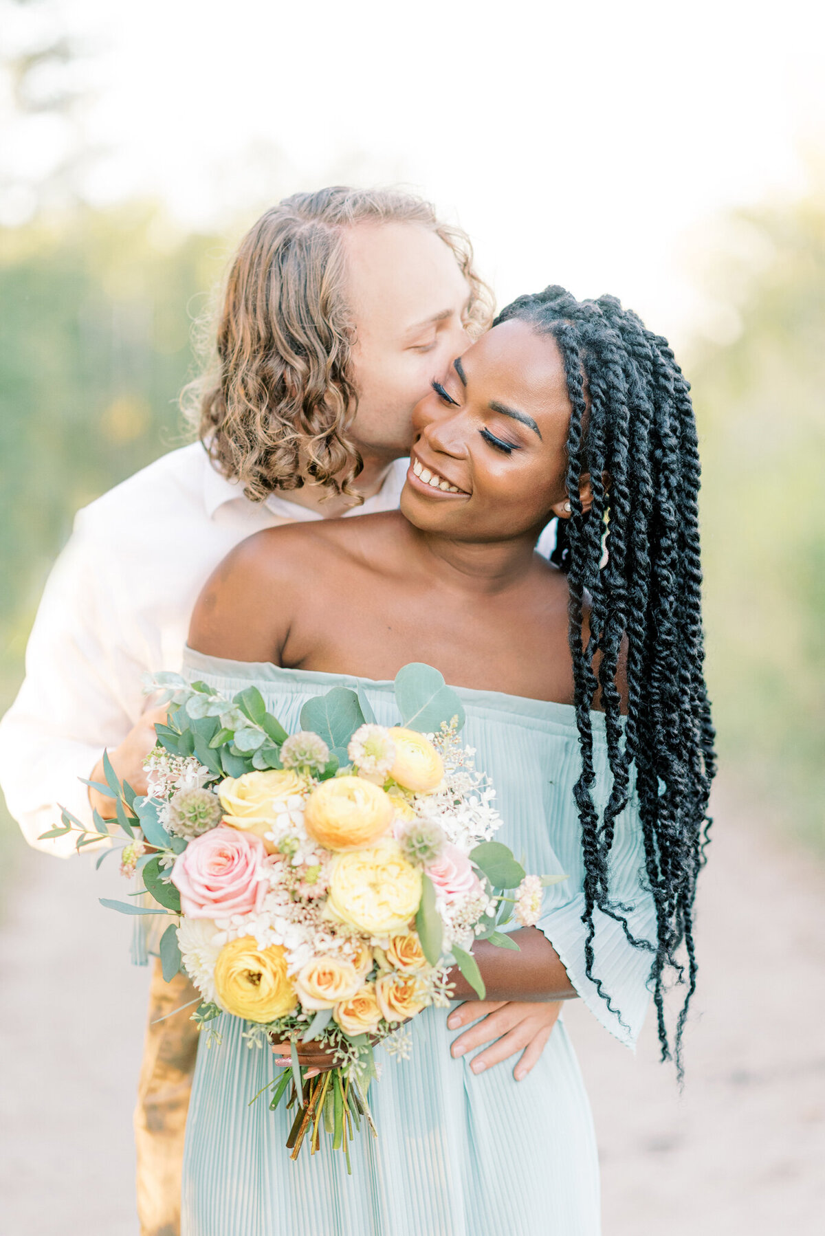 Classic, romantic interracial couple embracing, woman wearing a trendy pleated chiffon dress holding a stunning bouquet of pink and yellow spring flowers.