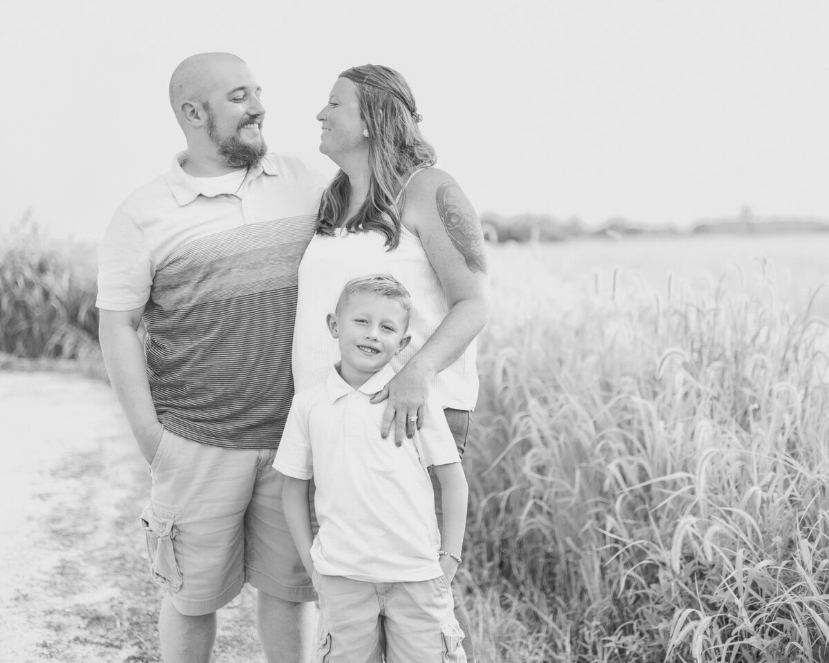Black and white photo of Mom, Dad, and son at a wildlife sanctuary.  Dad has shaved head with a beard, striped shirt, shorts and is looking at mom smiling.  Mom has long hair, wearing a sleeveless shirt, jeans, looking at dad smiling.  Son is wearing a short sleeve shirt, shorts, looking at the camera with a small smile and hands in his pockets
