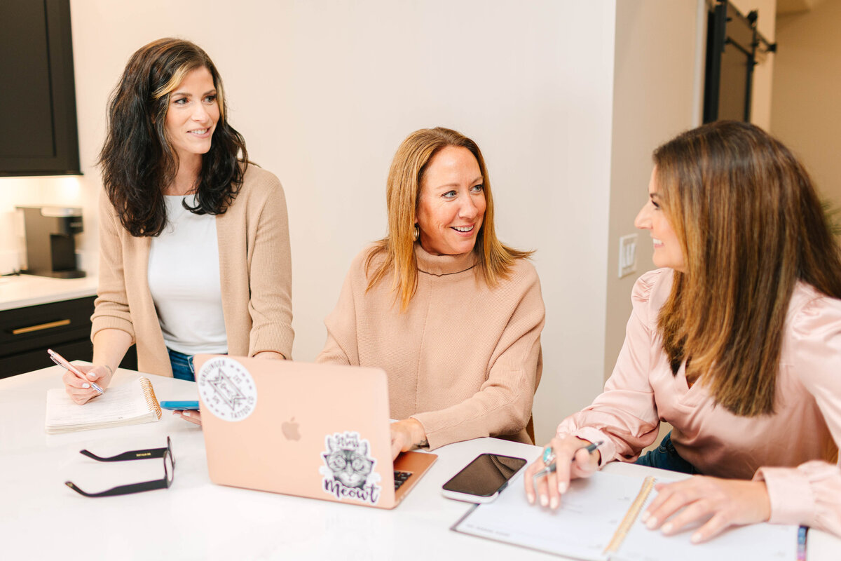 Three professional women collaborate at a white kitchen island, engaged in conversation. One types on a rose gold laptop, another takes notes, and the third gestures while speaking. They are dressed in neutral and blush tones, with a modern kitchen in the background