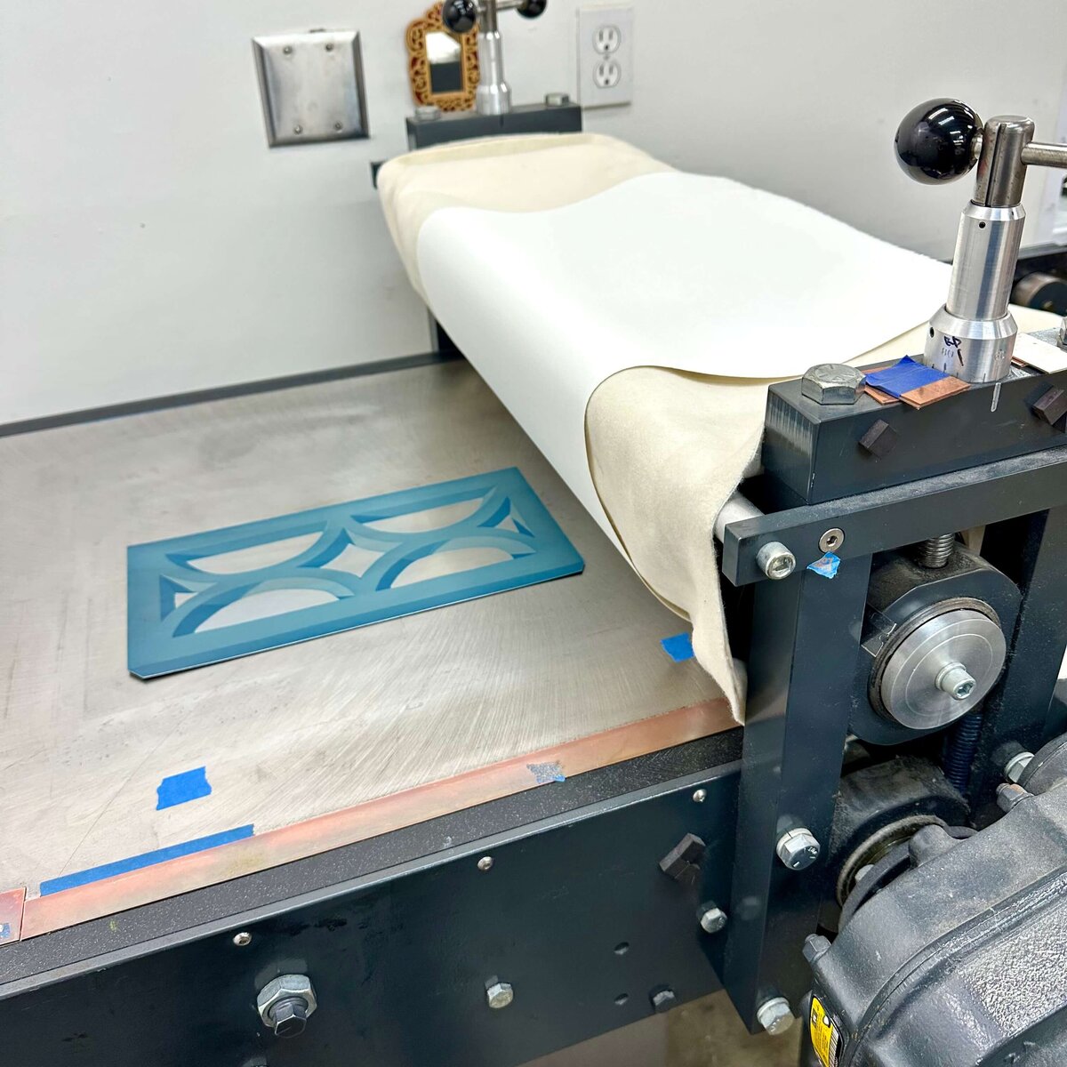 A light blue etched copper plate with intersecting arches is positioned on the etching press, ready for printing.