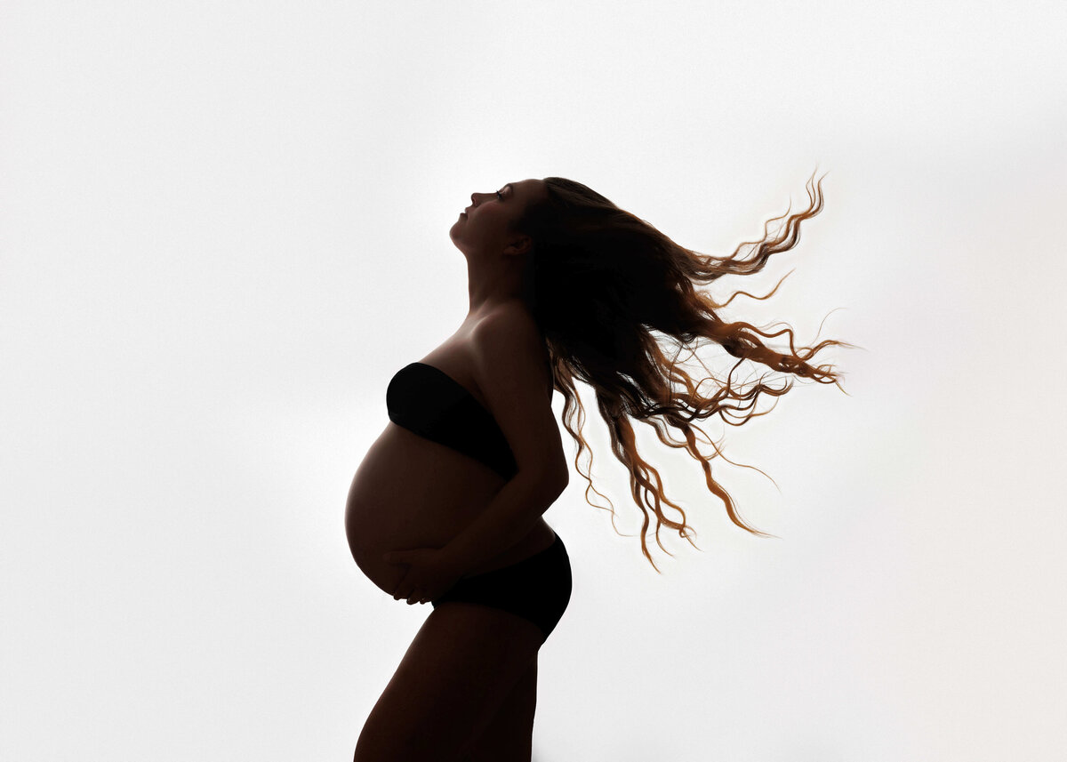 pregnant mother with her curly hair lying in the air wearing a black bandeu top with her pregnant stomch showing in front o a white  backlit curtain  with glitter in her hair at her maternity session in northern va