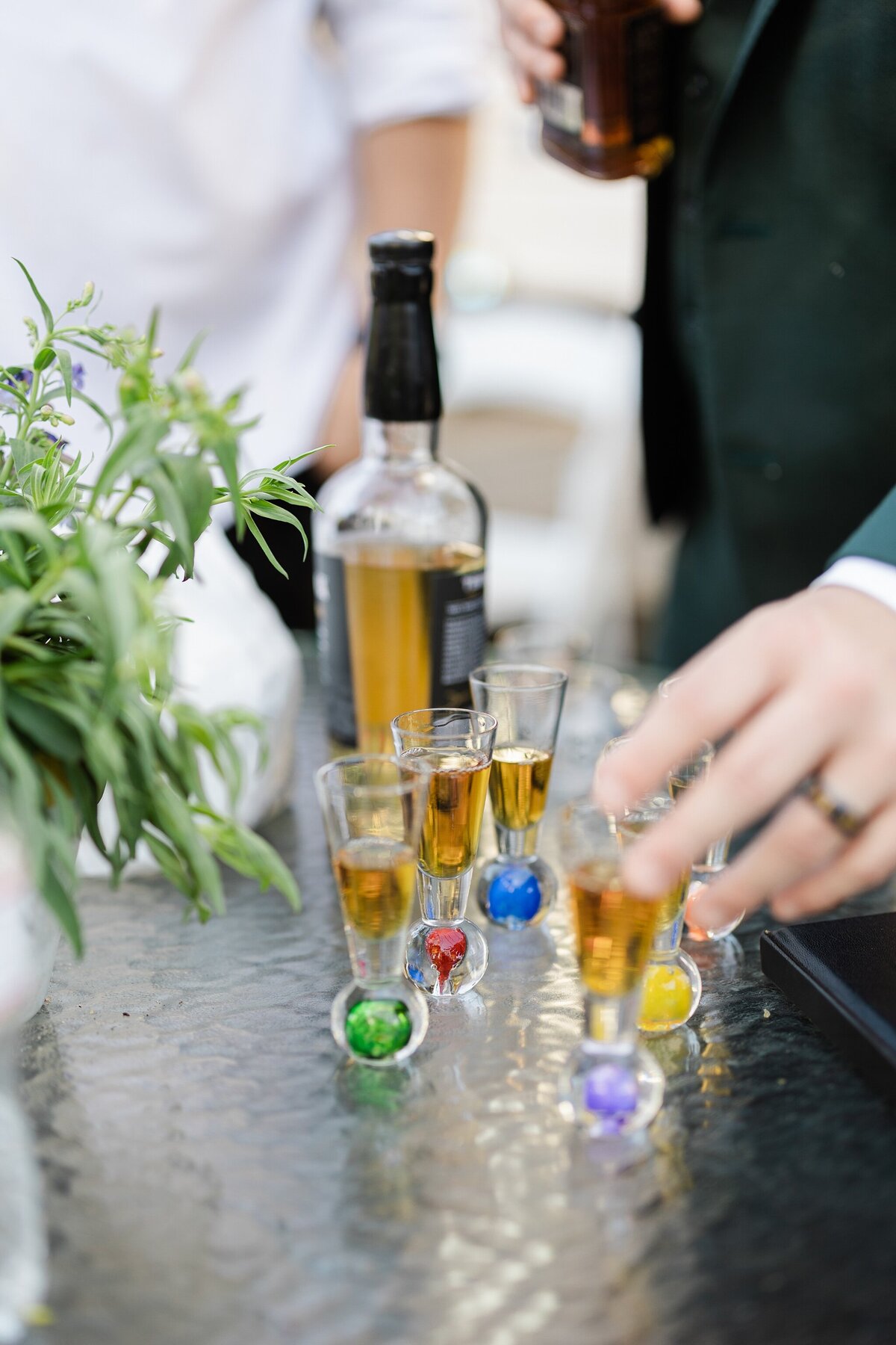 A detail shot of shot glasses being filled during a wedding reception in Dallas, Texas. Each shot glass is filled with dark liquor and is the bottom of each shot glass contains a glass sphere of a different color. A liquor bottle can be seen behind the group of shot glasses.