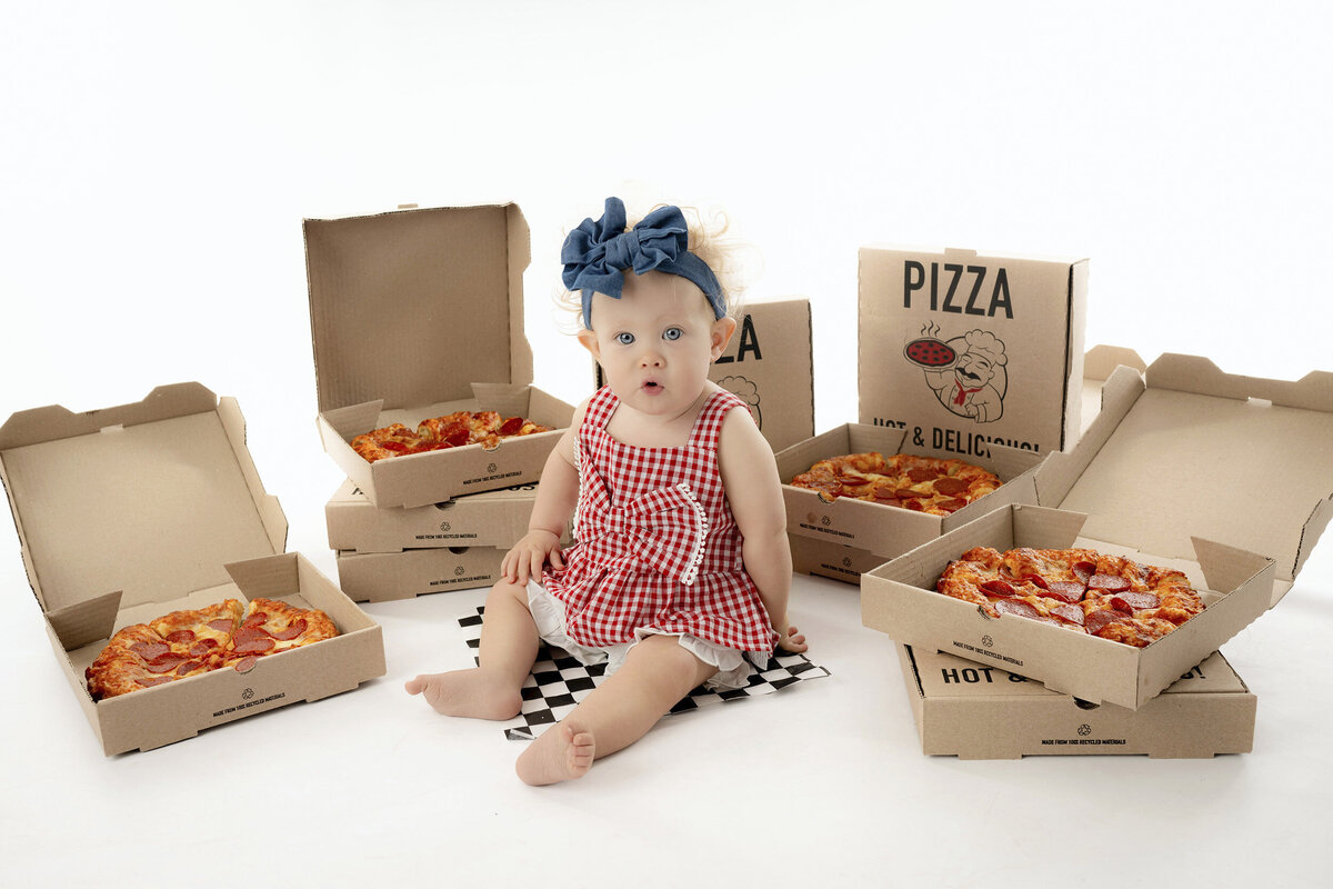 A toddler looks shocked in a red checkered dress sitting among many open pizza boxes.