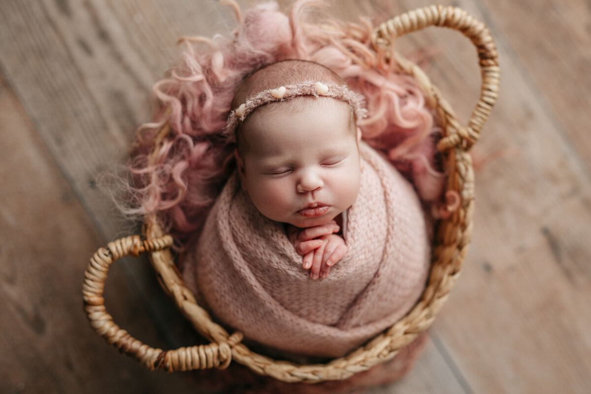 Studio newborn photography - baby sleeping in woven basket on wood floor. Baby is wrapped in a dusty light pink swaddle with delicate matching headband.