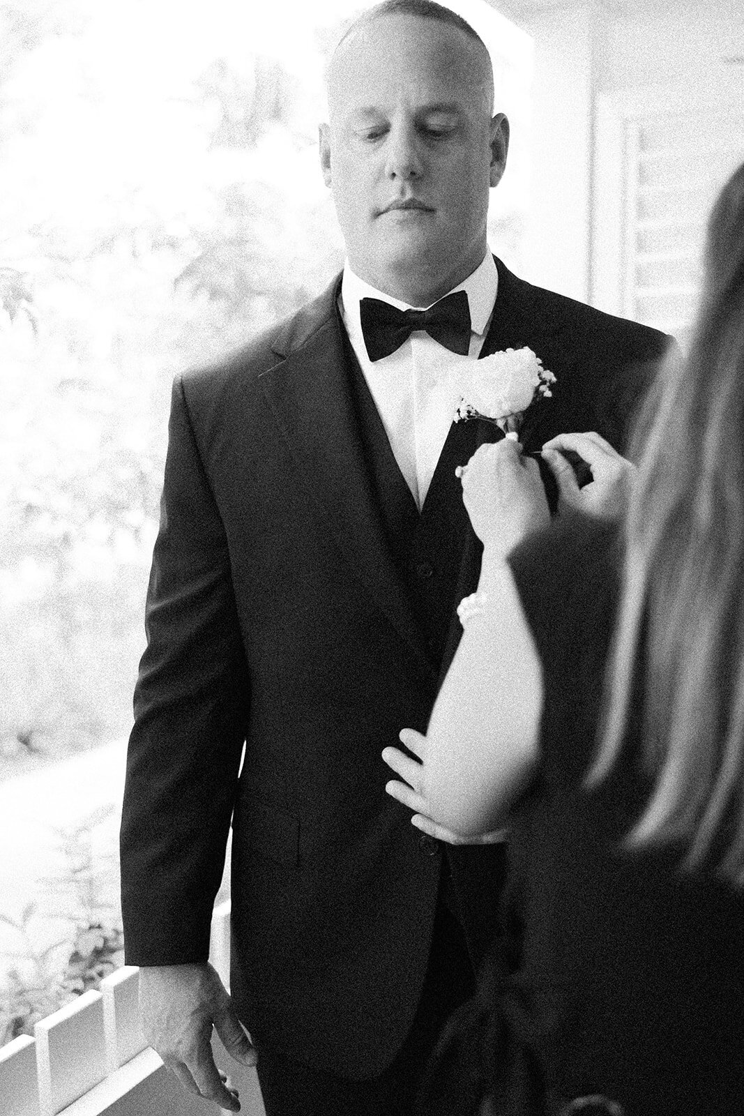 A touching black-and-white image of the groom’s mother carefully pinning a floral boutonniere on his lapel, capturing a sentimental wedding preparation moment. Photographed by Claudia Amalia Photography at Stephanie and Bradley’s wedding in Islamorada.