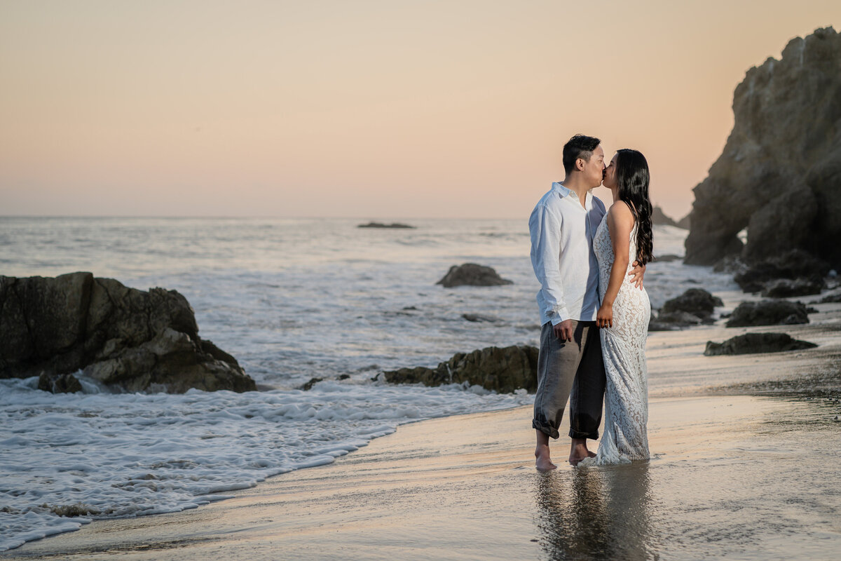 Asian couple eloping at El Matador State Beach in Malibu during low-tide sunset and kissing with rocky cliff beach.