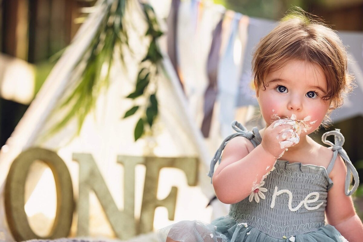 A baby girl, with a face covered in cake frosting, sits in front of a blurred backdrop featuring a teepee and the word "ONE" spelled out in wooden letters. The baby is dressed in a light blue dress with a white flower and the word "one" embroidered on it. She is wearing a big smile and looks delighted with the cake. The image captures the joy and messiness of a first birthday celebration.