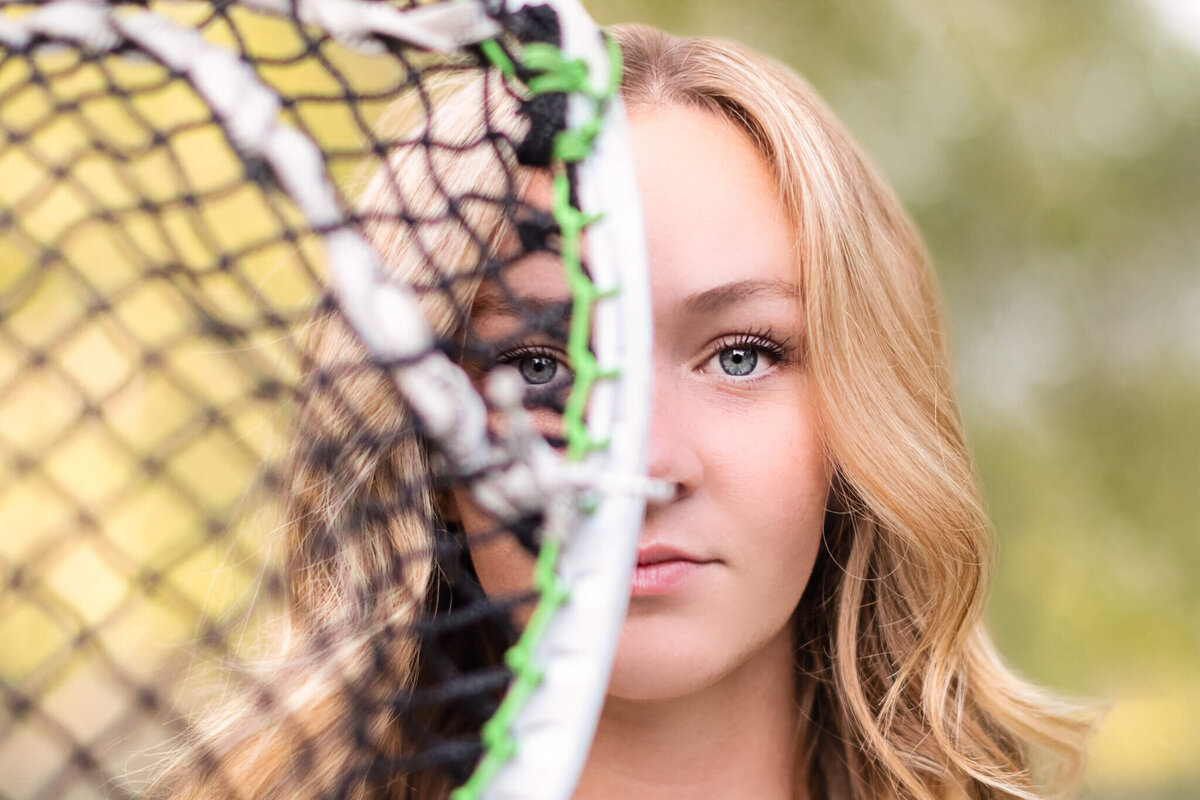A close-up of a young woman with blonde hair and blue eyes holding her lacrosse stick up to the camera, so the right side of her face is covered by the lacrosse net and the other side is in focus.