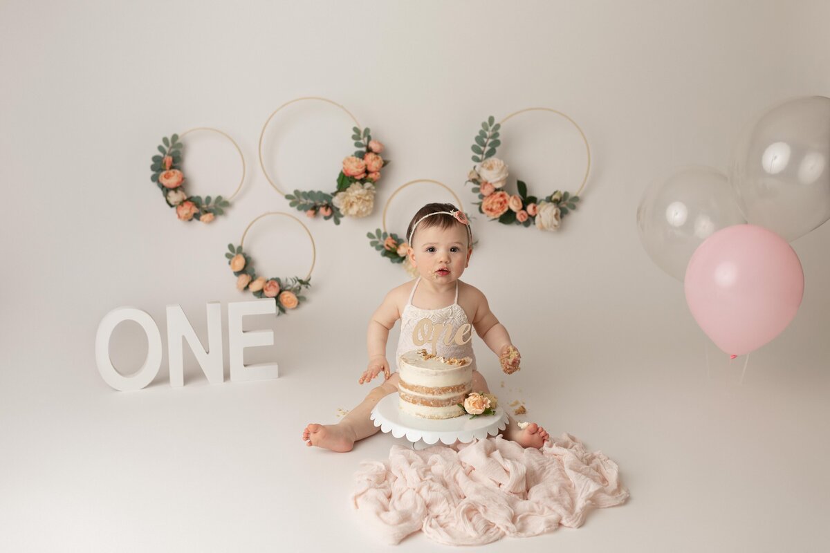 Cake Smash Photoshoot in London, Ontario Studio, pink, floral vibes. Baby is looking at camera and about to dig in to cake.
