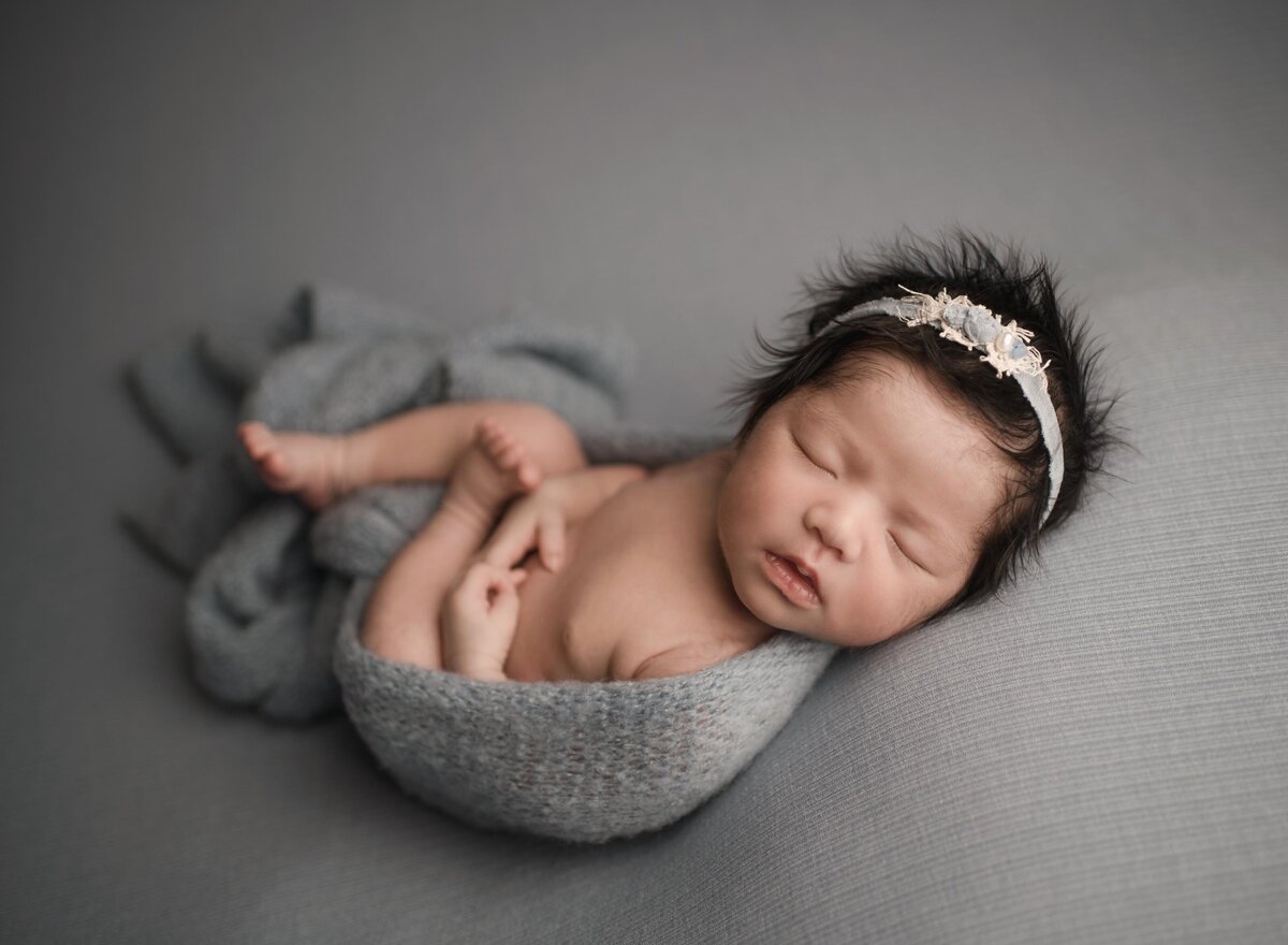 Baby girl is posed on her back for her newborn photoshoot with Lake Elsinore's best newborn photographer Bonny Lynn Photography. Baby has grey knit stretch fabric wrapped around her and is wearing a cream headband. Baby's head is turned so she is facing the camera.