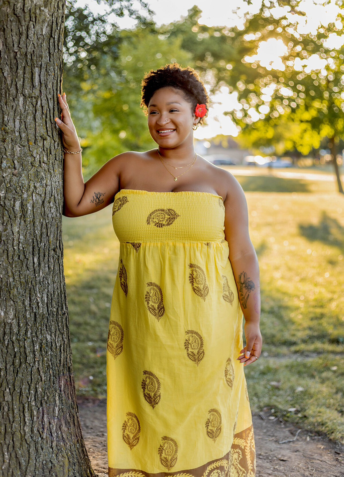 Woman in yellow halter sundress with red flower in her hair leaning on tree at sunset