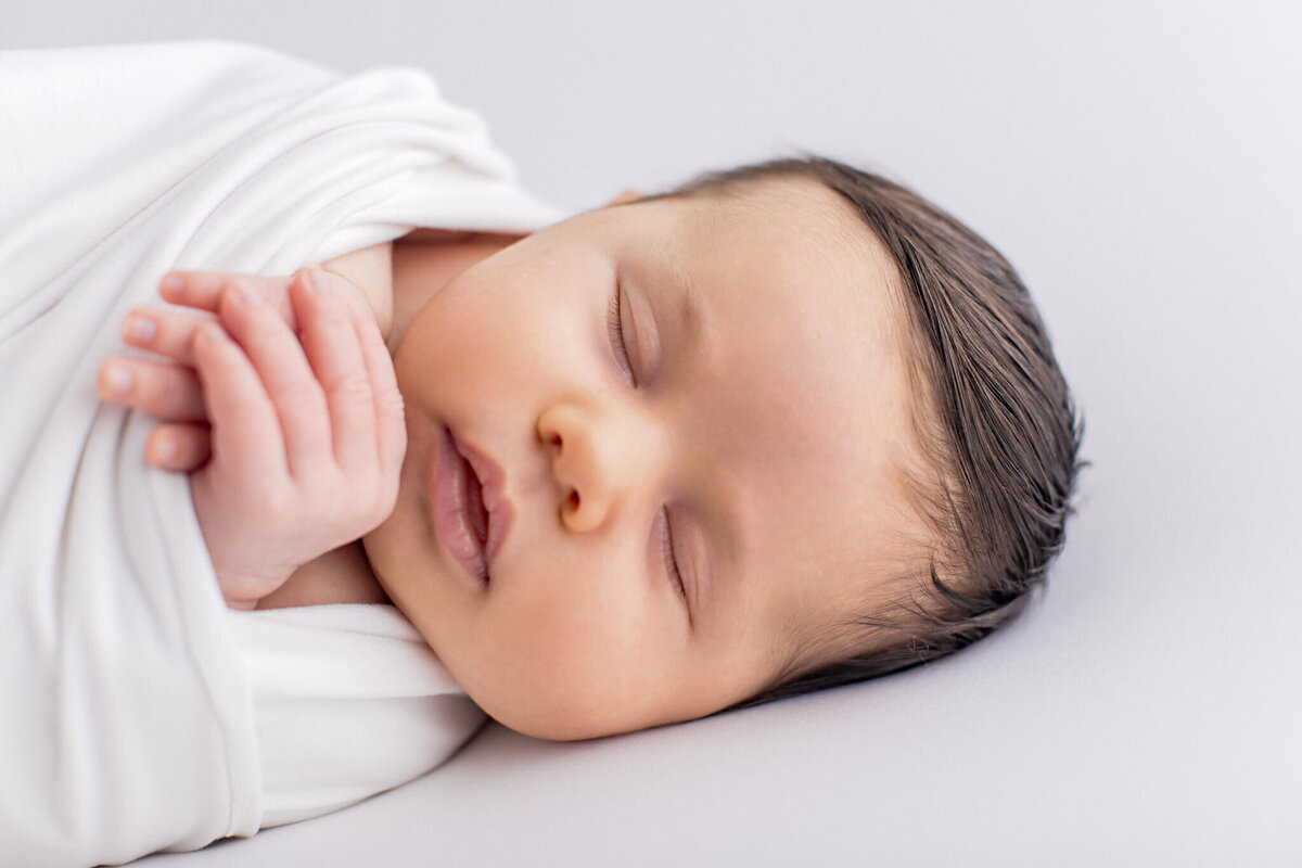 A peaceful, sleeping baby wrapped in a white blanket. The baby is lying on a soft, light-colored surface with eyes closed and hands gently clasped. The serene expression highlights the babys delicate features and calmness.