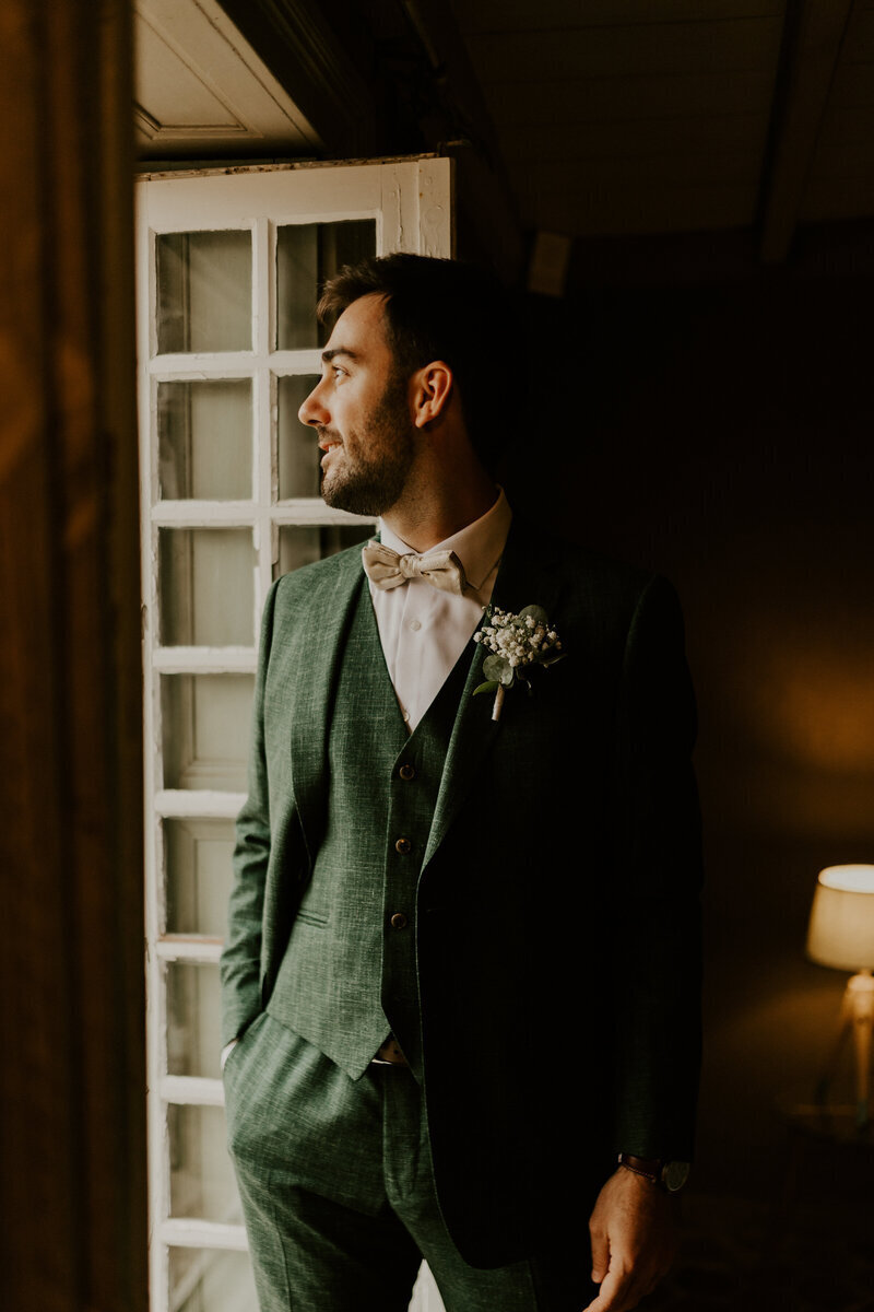 Marié en costume vert, mains dans les poches, regardant par la fenêtre ouverte. Ambiance tamisée immortalisée par laura, photographe de mariage en vendée.