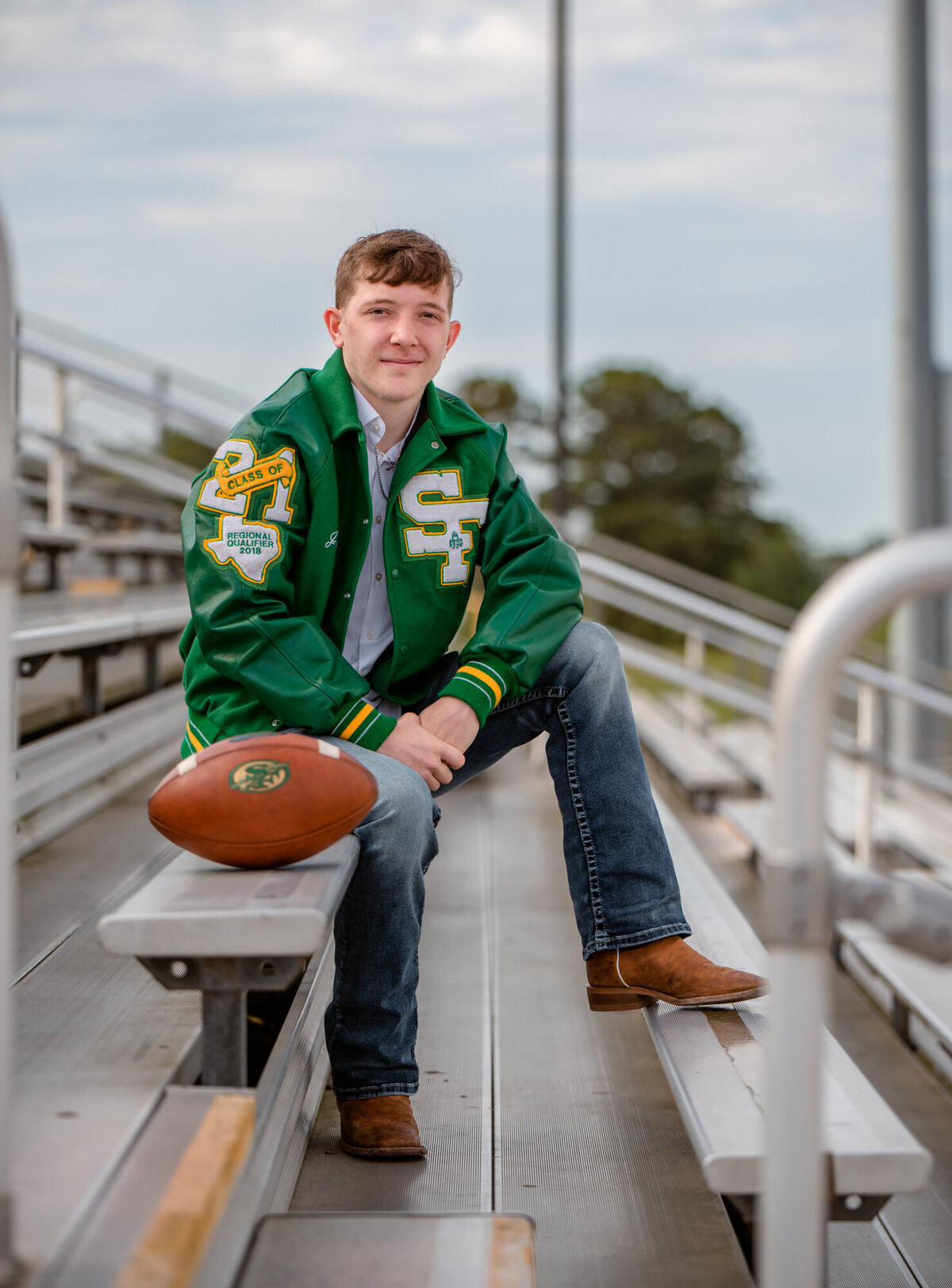 A senior boy sits in the football stands at his high school wearing his green letterman.