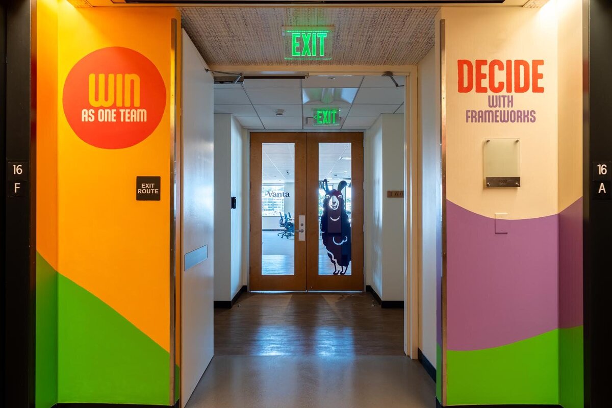 Office hallway with bright mural walls reading “Win as one team” and “Decide with frameworks,” leading to glass doors featuring a llama graphic.
