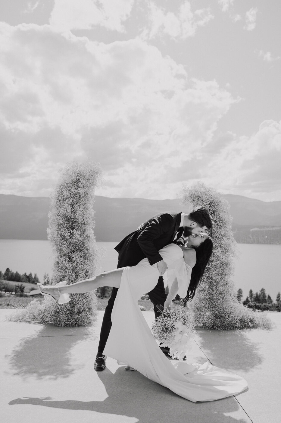 Kelowna Wedding Couple over looking the Okanagan Lake in Lake Country BC.  They are wearing modern sleek wedding attire with a gorgeous wedding dress and black tux.  A huge floral arch behind them filled with babysbreath.  A modern wedding day with a gorgeous look out!  They are dancing in their reception at the 50th Parallel Estate With a gorgeous classic black and white image