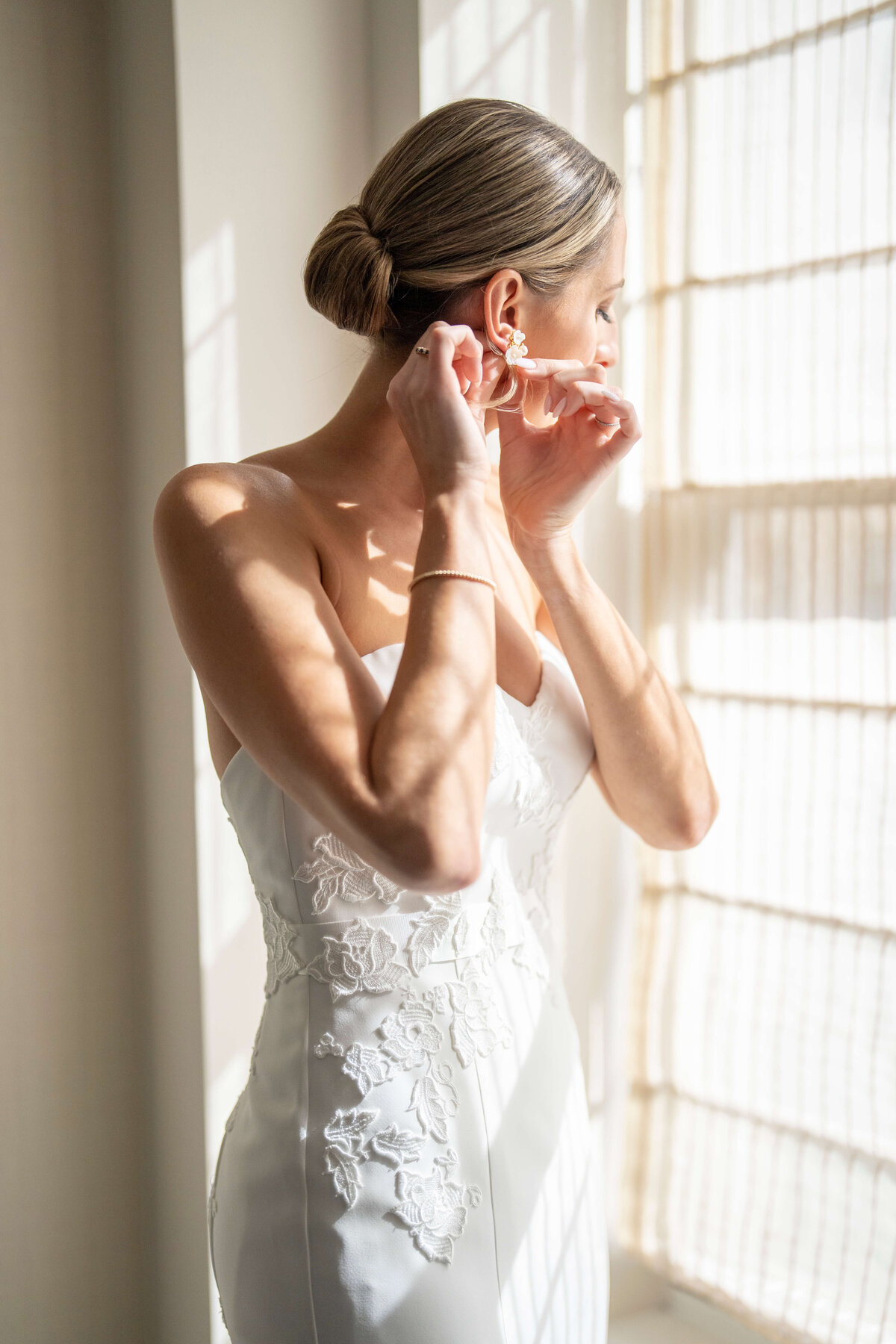A bride in a white dress stands near a window, adjusting an earring. Sunlight filters through sheer curtains, casting soft shadows. Her hair is elegantly styled in a bun.