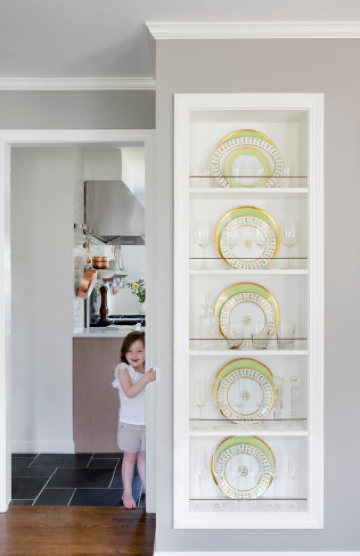 Custom shelves in the dining room display plates and china, with a view of the kitchen renovation. A family-friendly interior design in Westwood, Massachusetts.