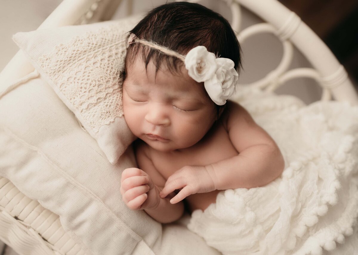 Side angled image. Baby sleeping on newborn  bed prop on cream bedding wearing a headband with flowers on the side.
