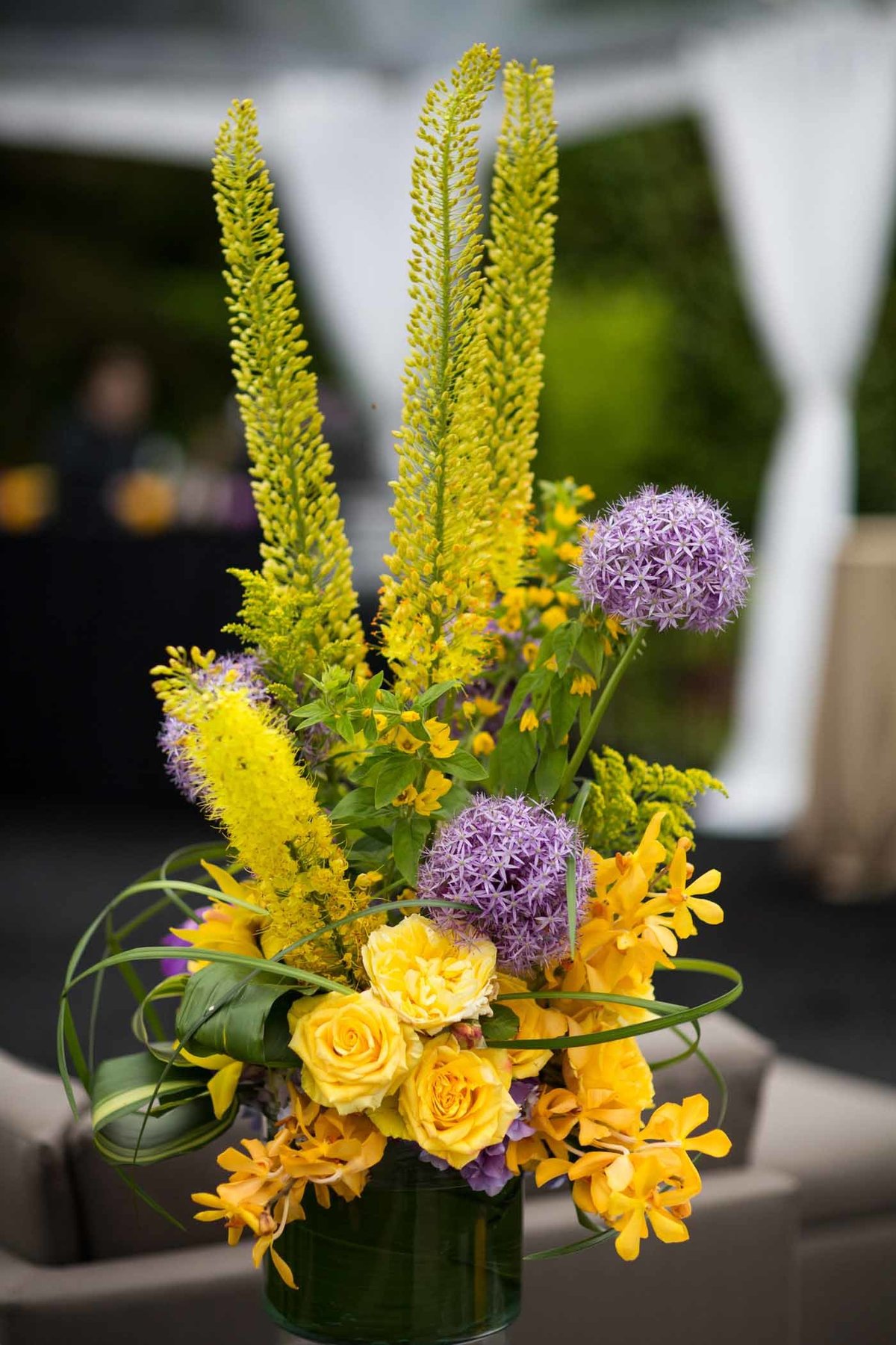 large flower arrangement of monstera leaves, large purple allium, yellow orchids, yellow eremurus, and yellow roses, with lily grass loops