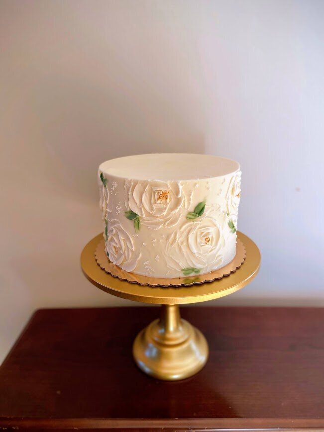 On a gold stand is shown a white cake with white pallet knife roses painted around the sides, accented with green leaves and gold centers.