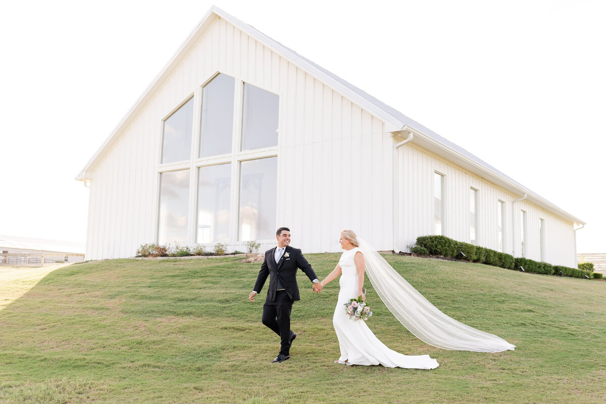 Groom leading Bride across grass while looking at each other in front of their wedding chapel at The Farmhouse