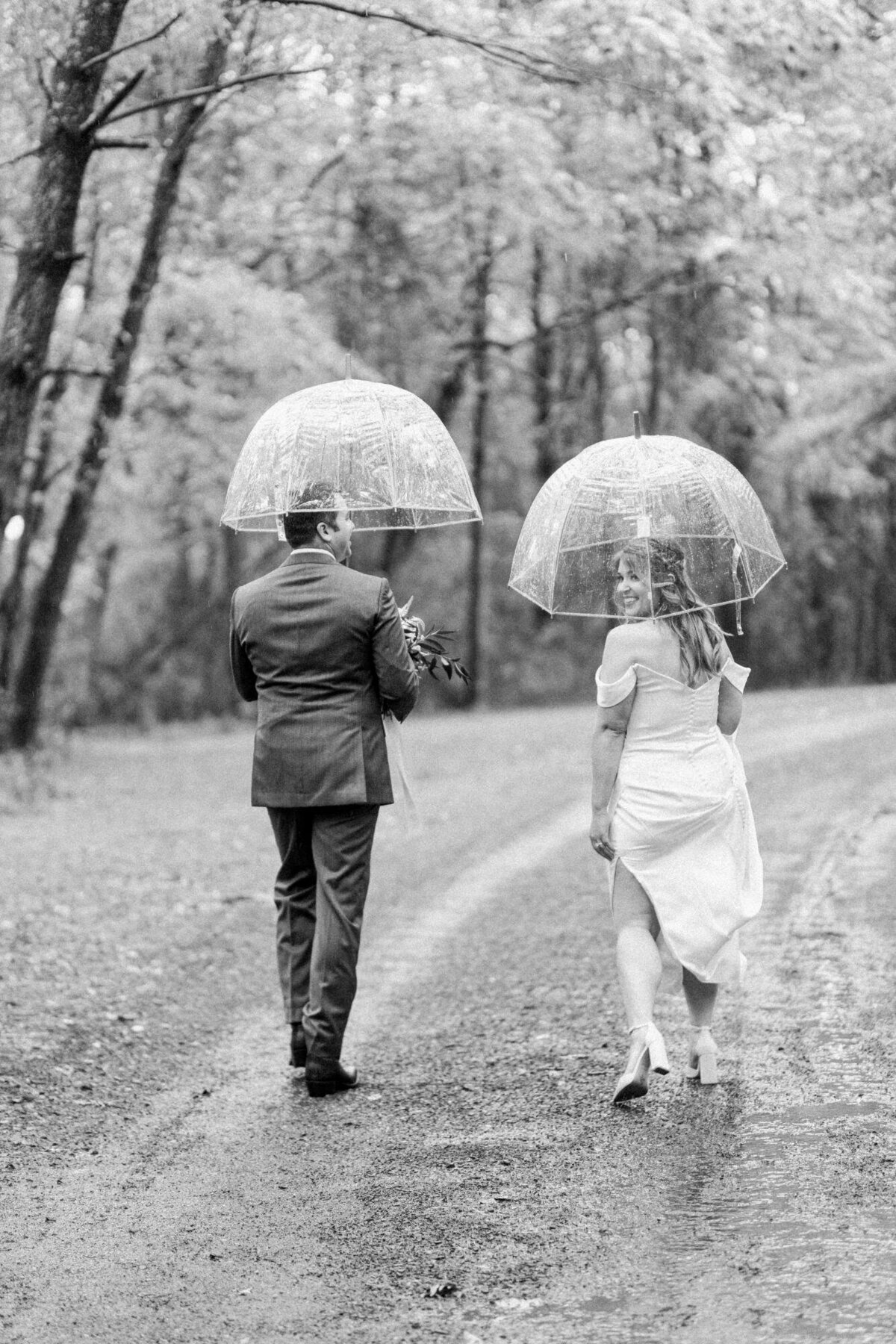 A couple walking under transparent umbrellas on a wet path with trees around, captured in black and white.