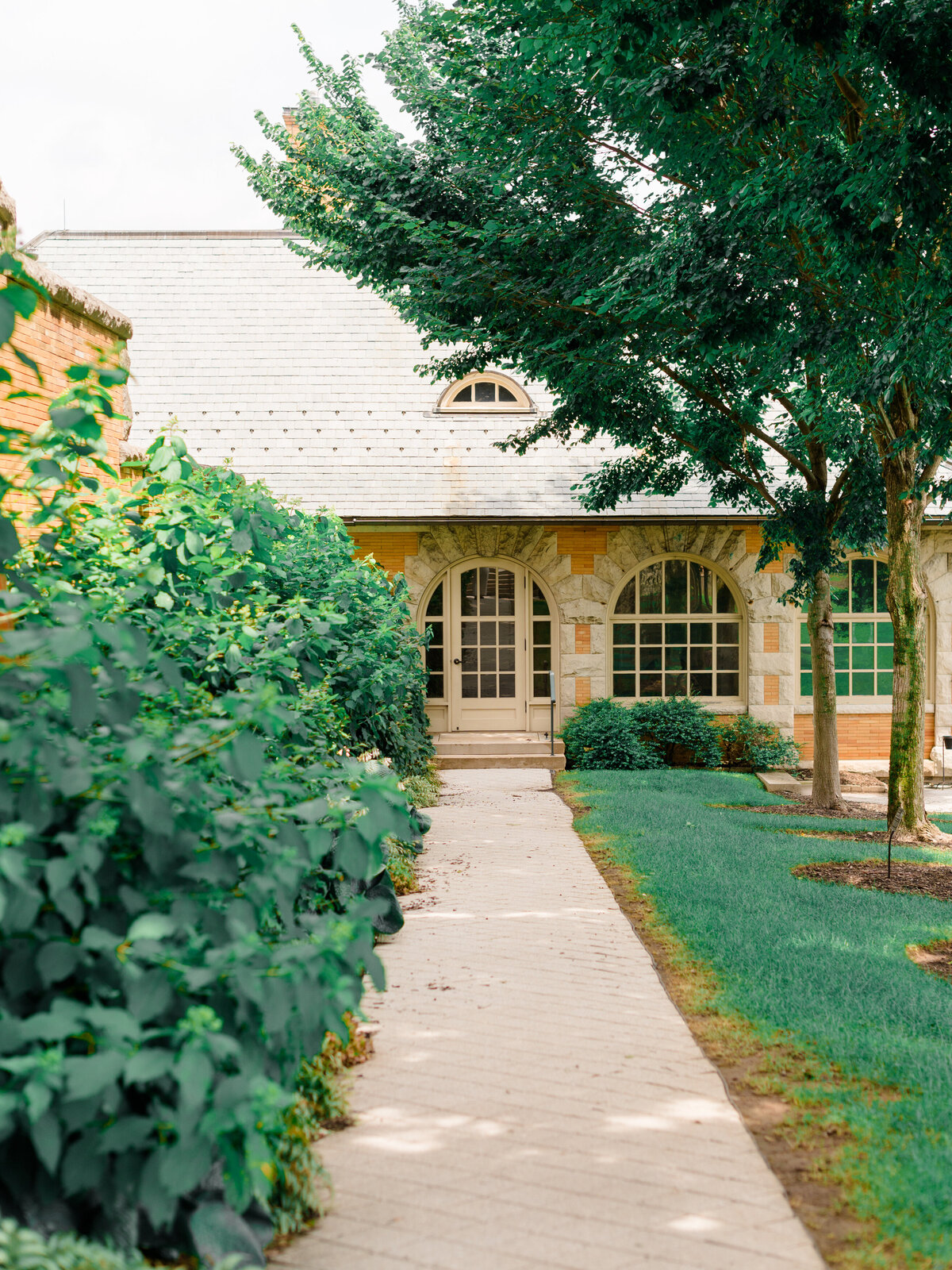 A charming garden pathway leads to the arched stone entryway of Cairnwood Estate, framed by manicured hedges and mature trees. This serene moment captures the timeless elegance of the venue, photographed by Elizabeth Kane Photography, a Luxury Philadelphia Wedding Photographer.
