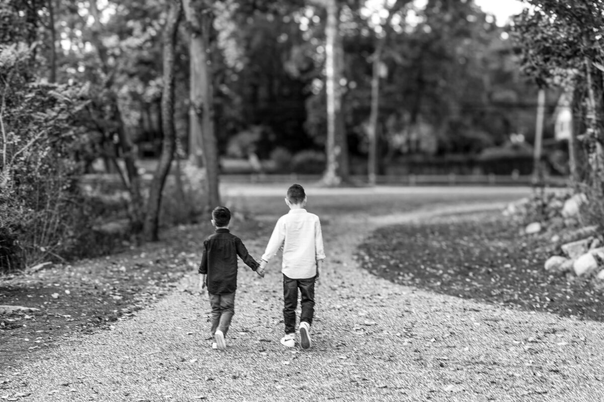 Black and white image of two young brothers walking away while holding hands along a path at Riverside Park in Connecticut.