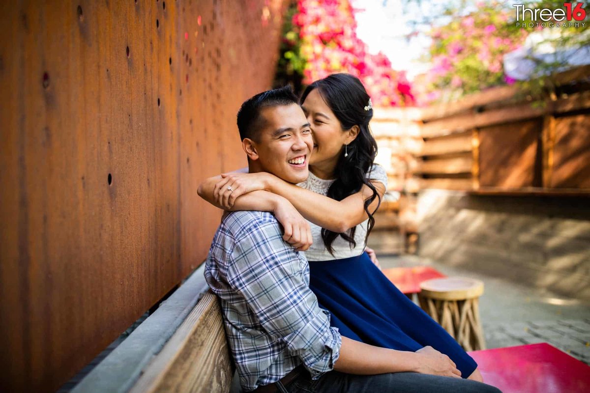 Bride to be sits on her fiance's lap and kisses his cheek