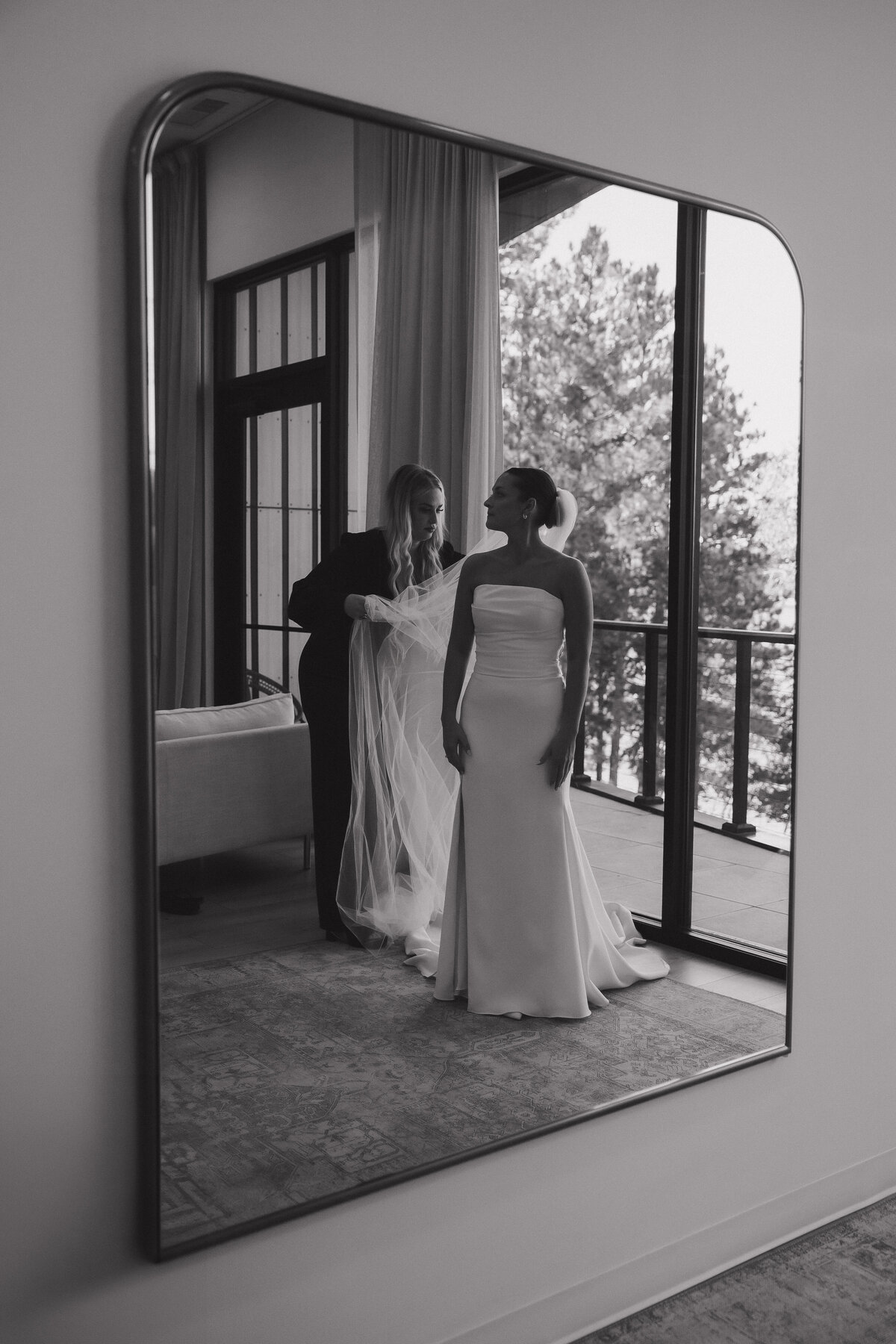 Through the reflection of a half-oval shaped mirror a woman in a black pant suit and platform sandals with long blond hair helps to extend a cathedral length veil out from behind a bride standing in a floor length wedding dress looking over her right shoulder with her hands at her side. The dress is white satin and strapless. There are pleats at the top of the dress creating texture to the dress and there is a thigh high leg slit. Behind the duo, there is a large window overlooking trees and a curtain that is the length of the wall. 