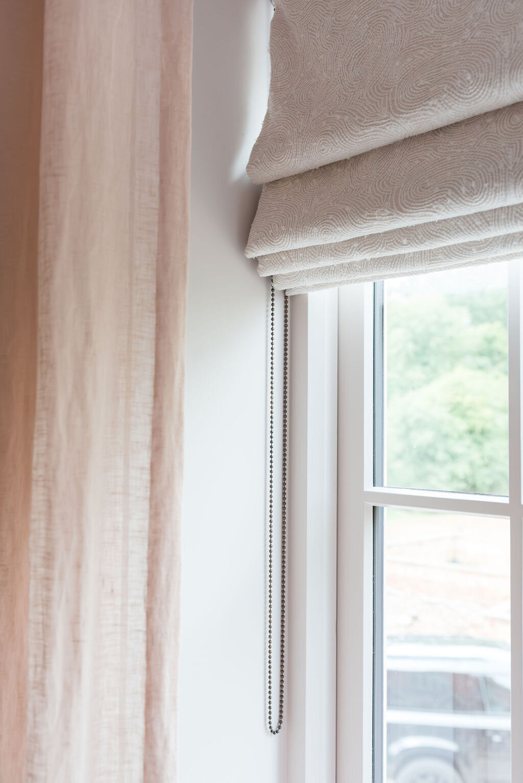 A close-up view of a window in a girl's bedroom with neutral textured blinds and soft pink drapes.