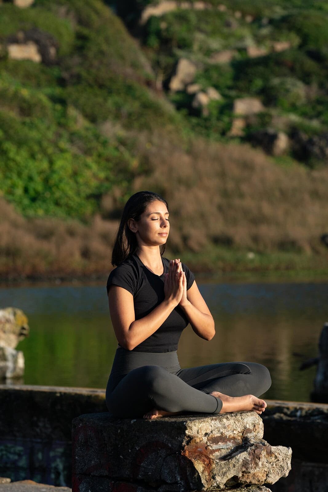 Woman sitting cross-legged, palms together, and closed eyes on a wall next to a pond at sunset