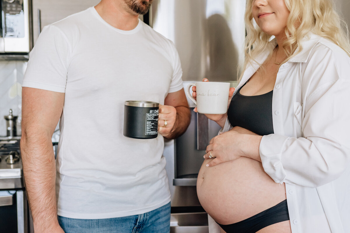 A close-up of the couple standing in the kitchen, focusing on their hands holding mugs. The man's mug says "#1 Dad," while the woman's mug reads "mama bear."