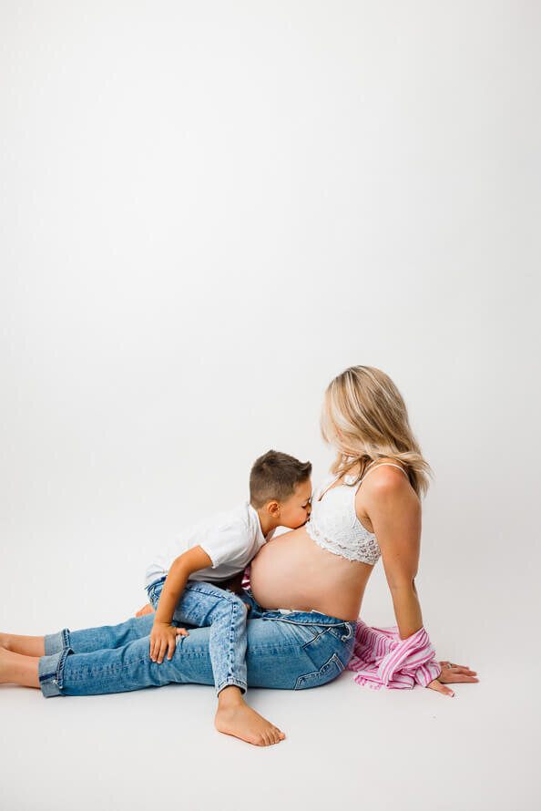 A pregnant woman sits on the ground while her son sits on her lap and kisses her belly.