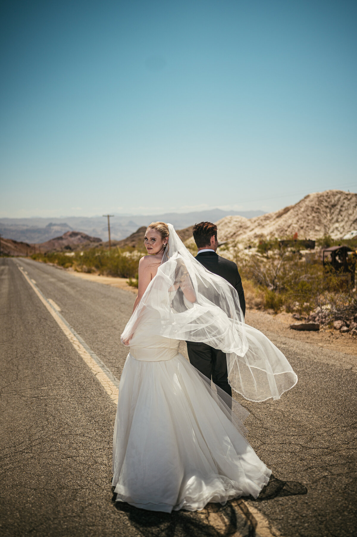 bride looking over her shoulder at photographer as she walks down the highway with new husband groom ELDORADO CANYON MINE TOURS nelsons ghost town weddings nelsons landing bride in sleeveless gown and cathedral veil  groom in black tux armani  las vegas elopements las vegas wedding photography las vegas wedding photographers mk delacy photography