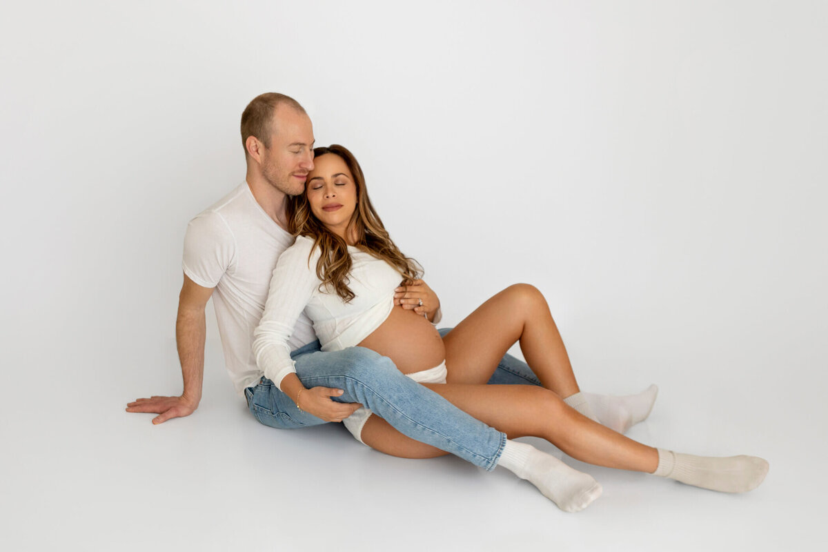 A pregnant woman reclines against her partner on a white studio floor. She is dressed in a white long-sleeve top, underwear, and socks, with her bare baby bump prominently visible. Her partner, wearing a white t-shirt and jeans, sits behind her, gently resting his head against hers with a serene expression. The woman has her eyes closed and one hand resting on her belly, creating a peaceful and intimate moment. The clean, minimalist background accentuates the couple's connection and the anticipation of their growing family.