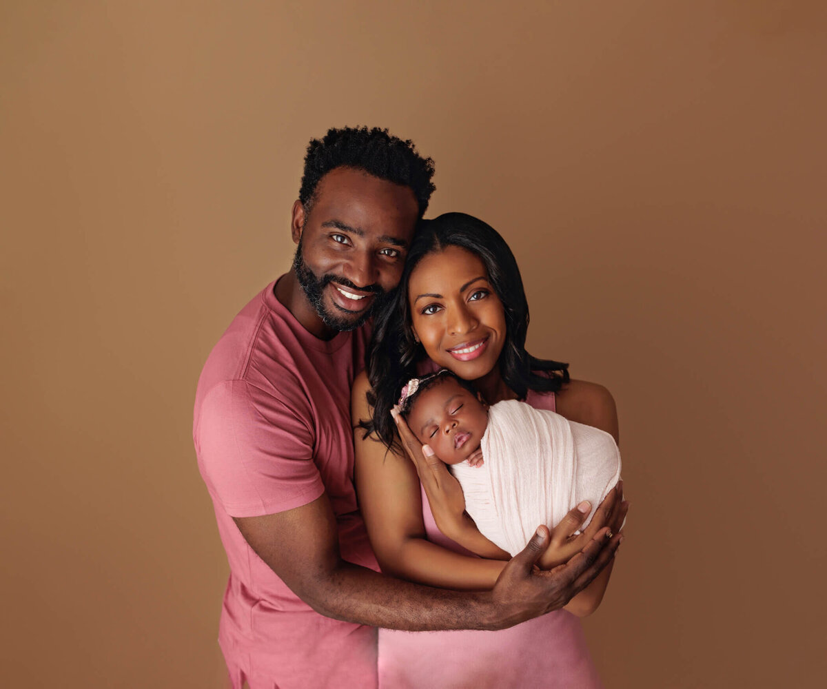 A joyful family portrait of a smiling couple holding their newborn baby girl. The mother and father are both dressed in matching shades of pink, with the baby swaddled in a white blanket and wearing a floral headband. They stand against a warm brown background.