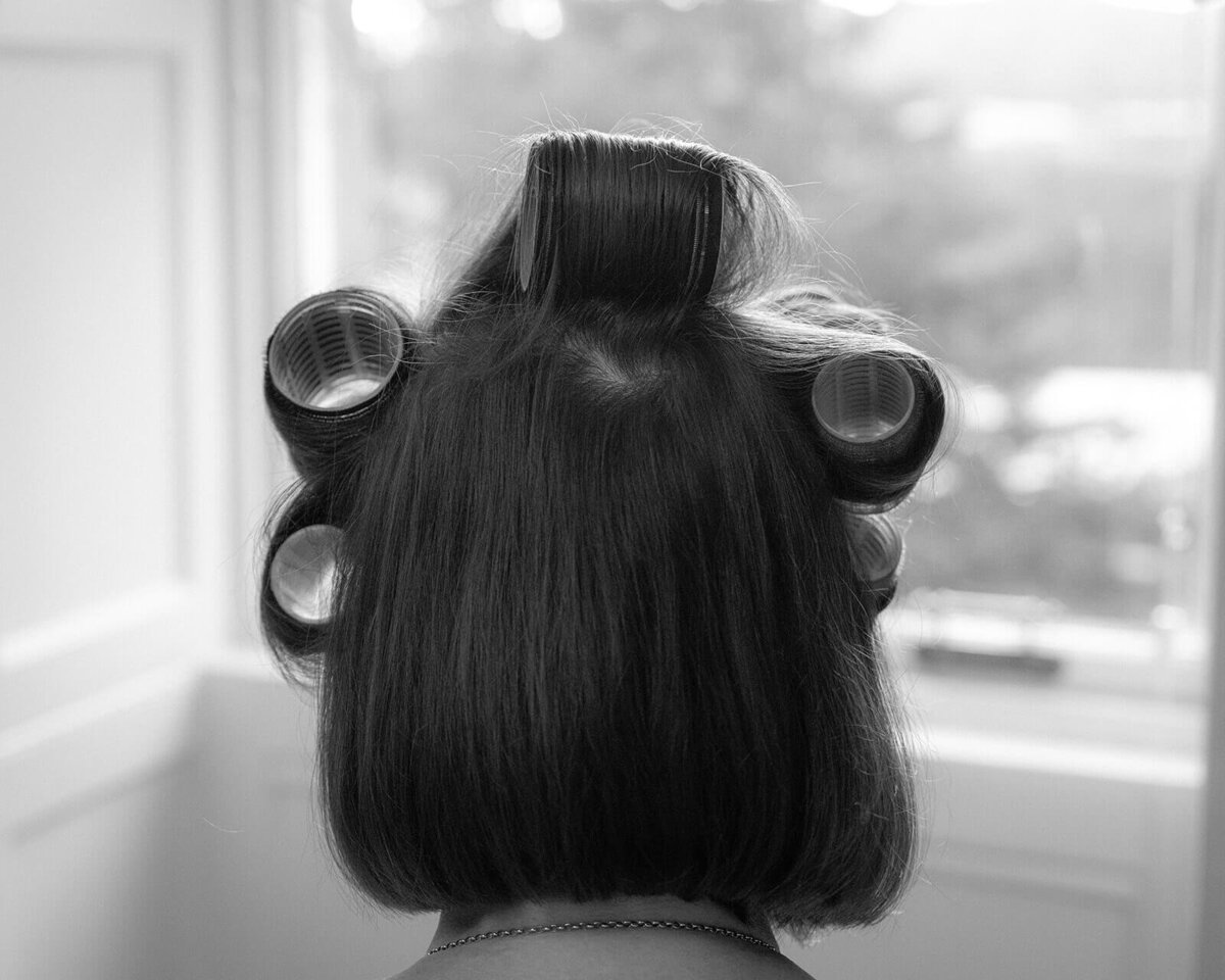 A mother of the bride faces a window with 5 curlers in her hair. The image is focussed on her head only