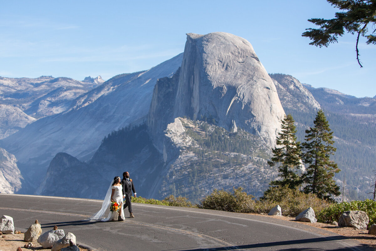 A bride and groom walk along a road in yosemite with Half Dome in the background.