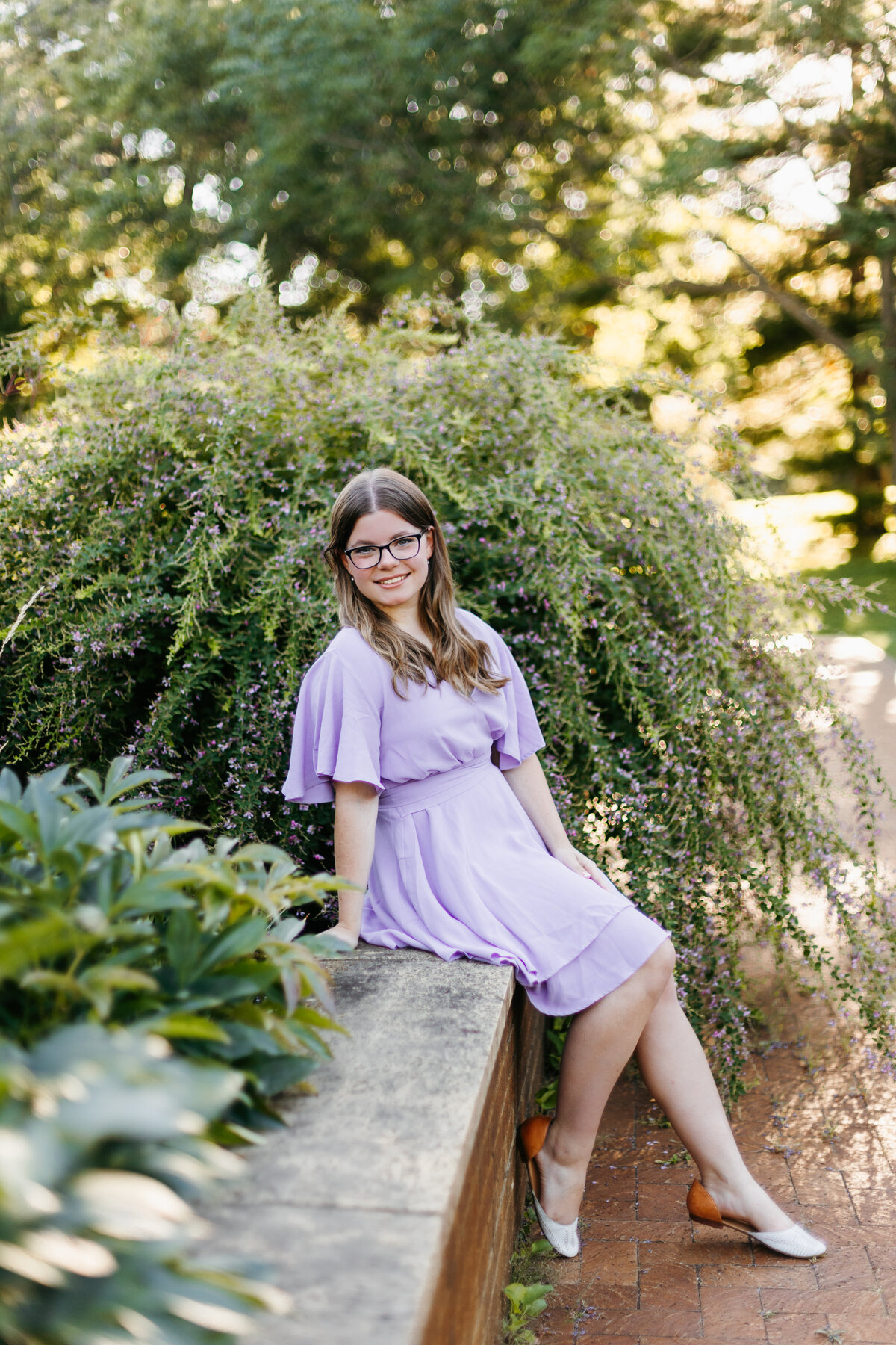 Senior girl in a light purple dress sitting on a short wall in front of a large bush