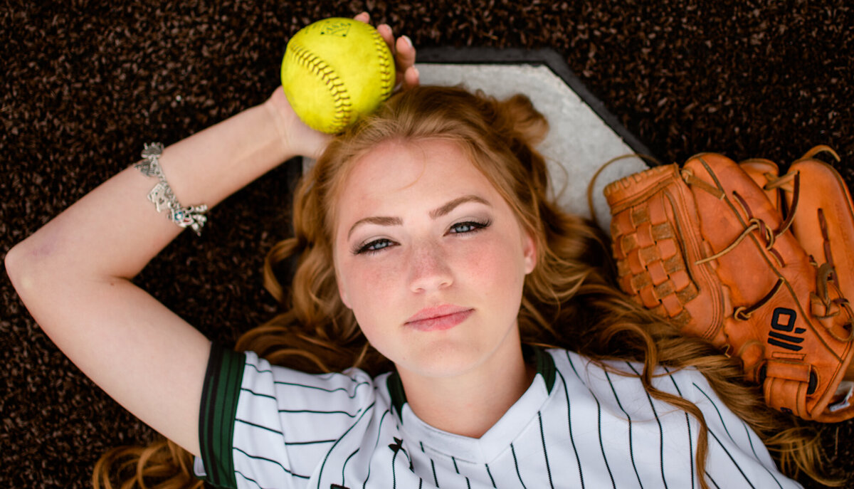 A senior softball player lays at home plate with her glove while holding a softball.