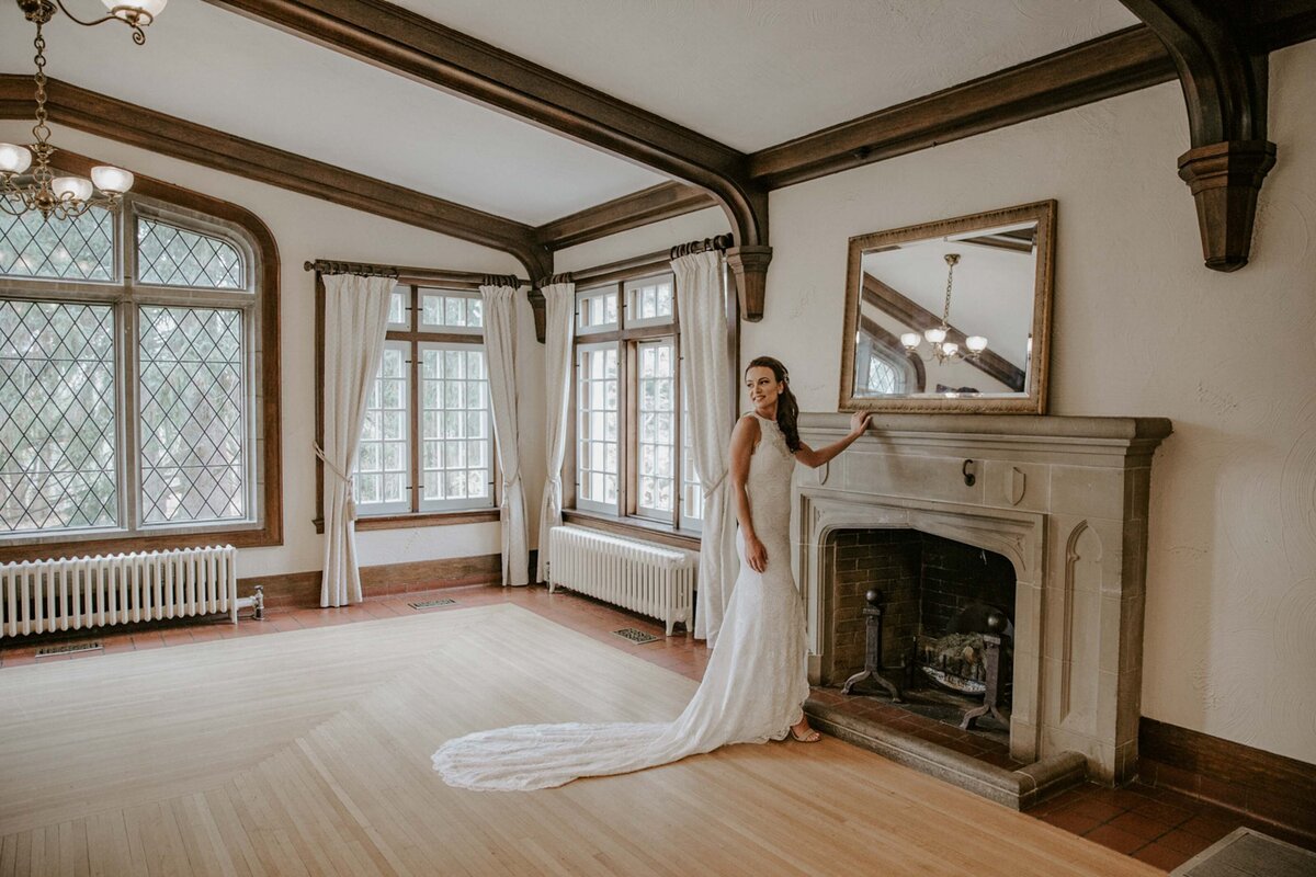 Bride standing in front of tall stone fireplace at Elsie Perrin Williams Estate in London, Ontario. Bride is gently resting her hand on the mantle and looking over her shoulder to the distance.