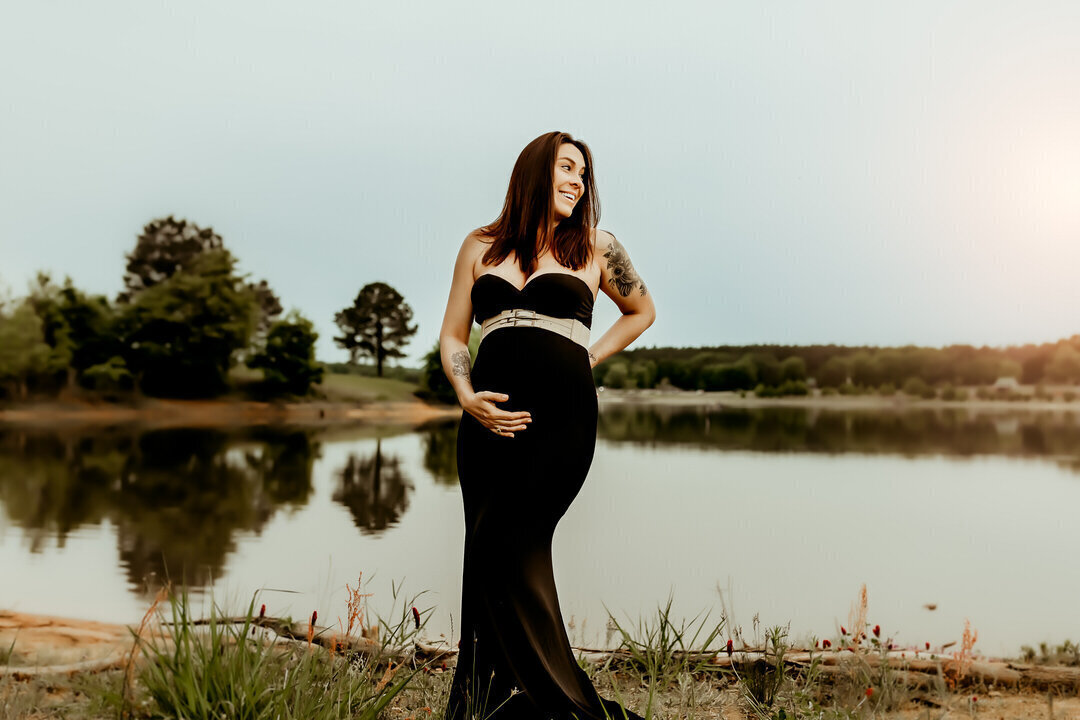Pregnant woman in a black dress stands by a lake, smiling and holding her belly. Trees and calm water are in the background.