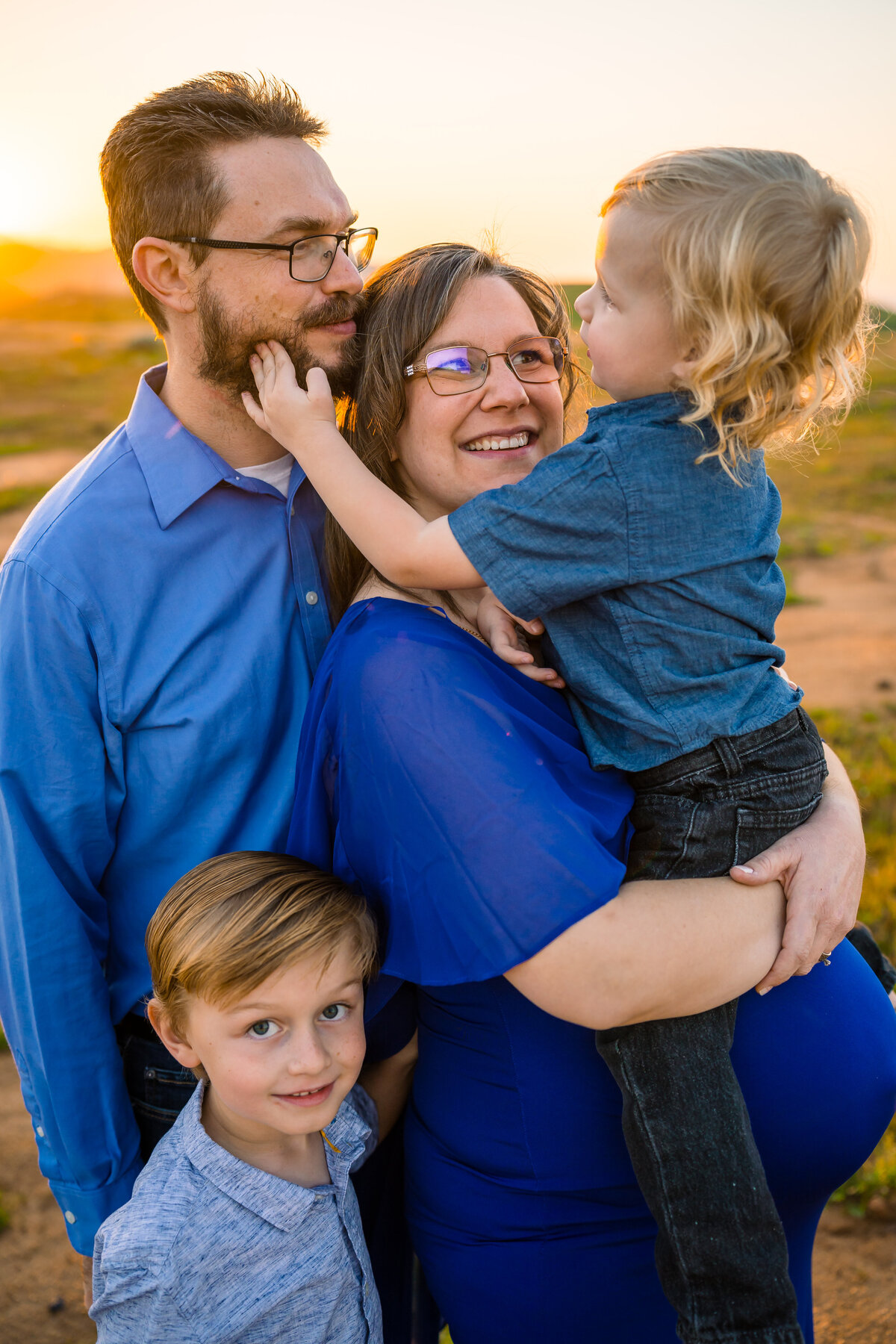 A family, dressed in blue, stands closely together on a hilltop at sunset, and the blonde toddler sitting on mom's pregnant belly reaches behind her to touch his dad's beard while his brother looks up and smiles at the camera. Photo by SAVI Photography - Los Angeles CA Photographer