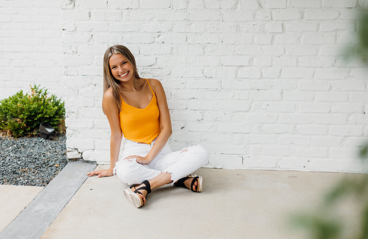 Senior girl in an orange top and white pants sitting on the ground in front of a white brick wall